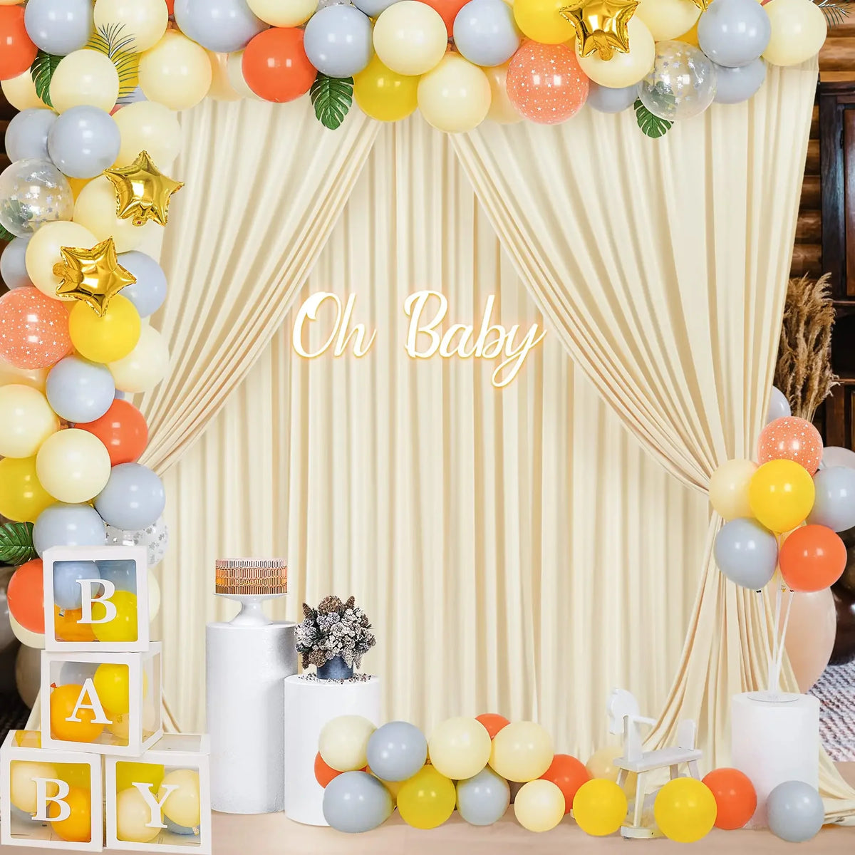 A decorative backdrop featuring a cream curtain draped with clusters of yellow, orange, and gray balloons for a baby theme.