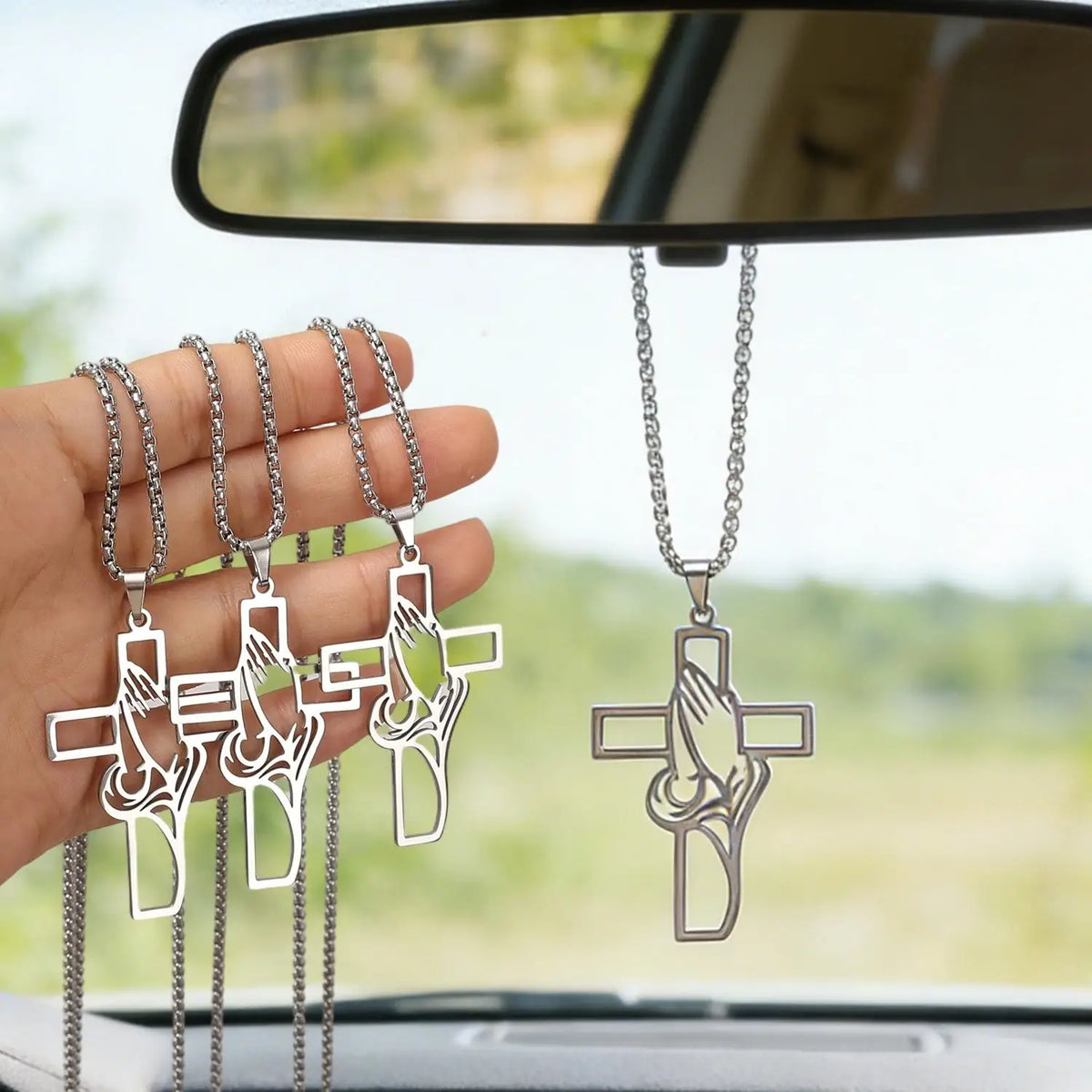 Stainless steel praying hands cross pendant hanging from a chain displayed in front of a car mirror.