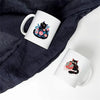 A white mug featuring a black cat design with pink and blue flowers, placed on a dark blue fabric.
