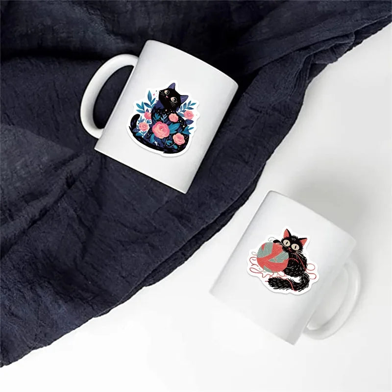 A white mug featuring a black cat design with pink and blue flowers, placed on a dark blue fabric.