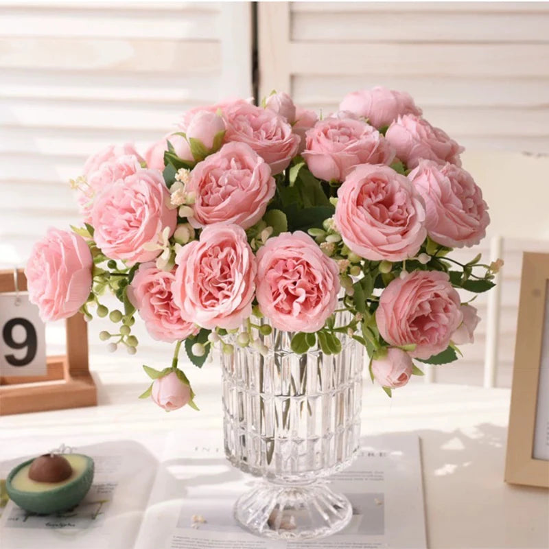 A bouquet of pink silk peony roses displayed in a clear glass vase on a white surface.