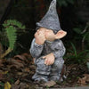 A small statue of a gnome with a pointed hat and gray beard sitting in a garden setting.