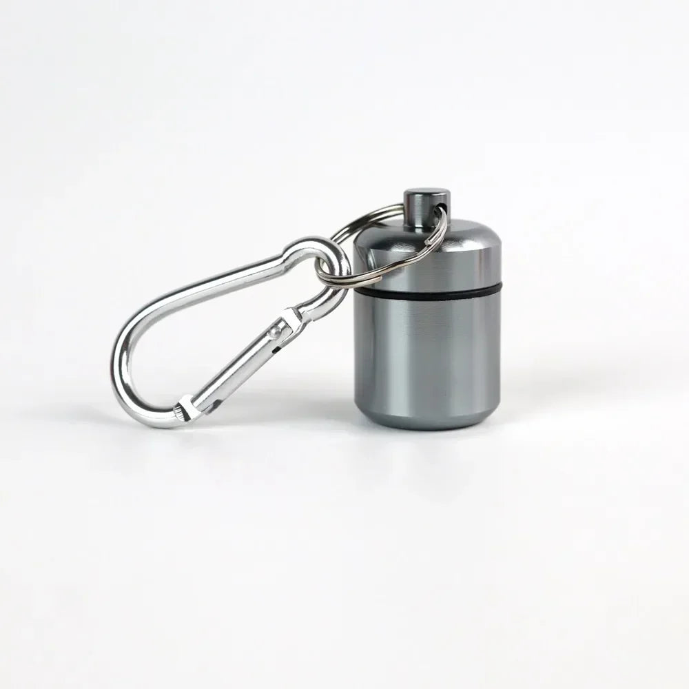 A small, silver pill organizer is attached to a keyring and presented against a white background.