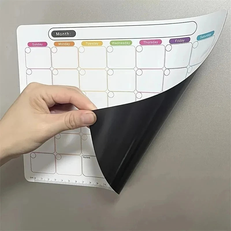 A hand holds a magnetic fridge planner with monthly grids and erasable notes on a gray wall.
