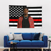 A man wearing a black cap and a black shirt stands in front of an American flag tapestry featuring stars and stripes.