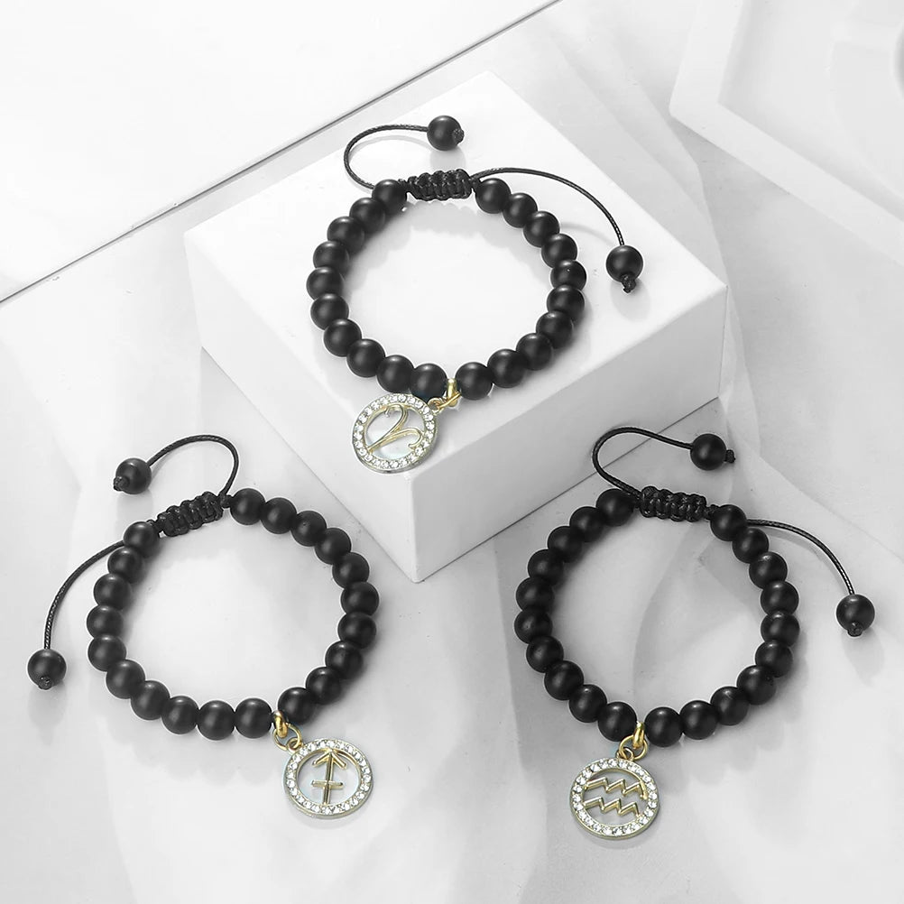 A black beaded bracelet with a gold charm featuring an astrological symbol is displayed on a white box.