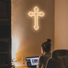 A glowing white neon cross wall sign is mounted on a light-colored wall, viewed from the side.