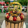 A round cake decorated with fresh berries and chocolate frosting, topped with a gold acrylic Happy Birthday topper.