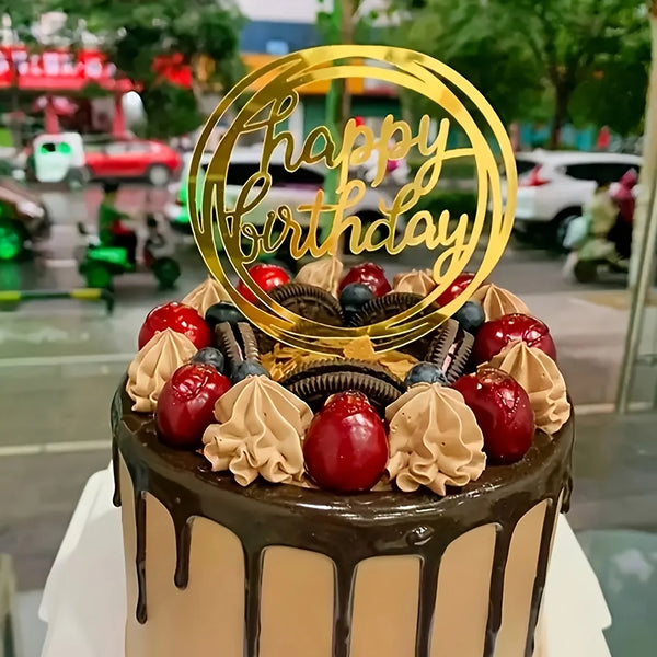 A round cake decorated with fresh berries and chocolate frosting, topped with a gold acrylic Happy Birthday topper.
