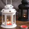 A white vintage hanging metal lantern with a glass enclosure and a lit candle on a wooden surface.