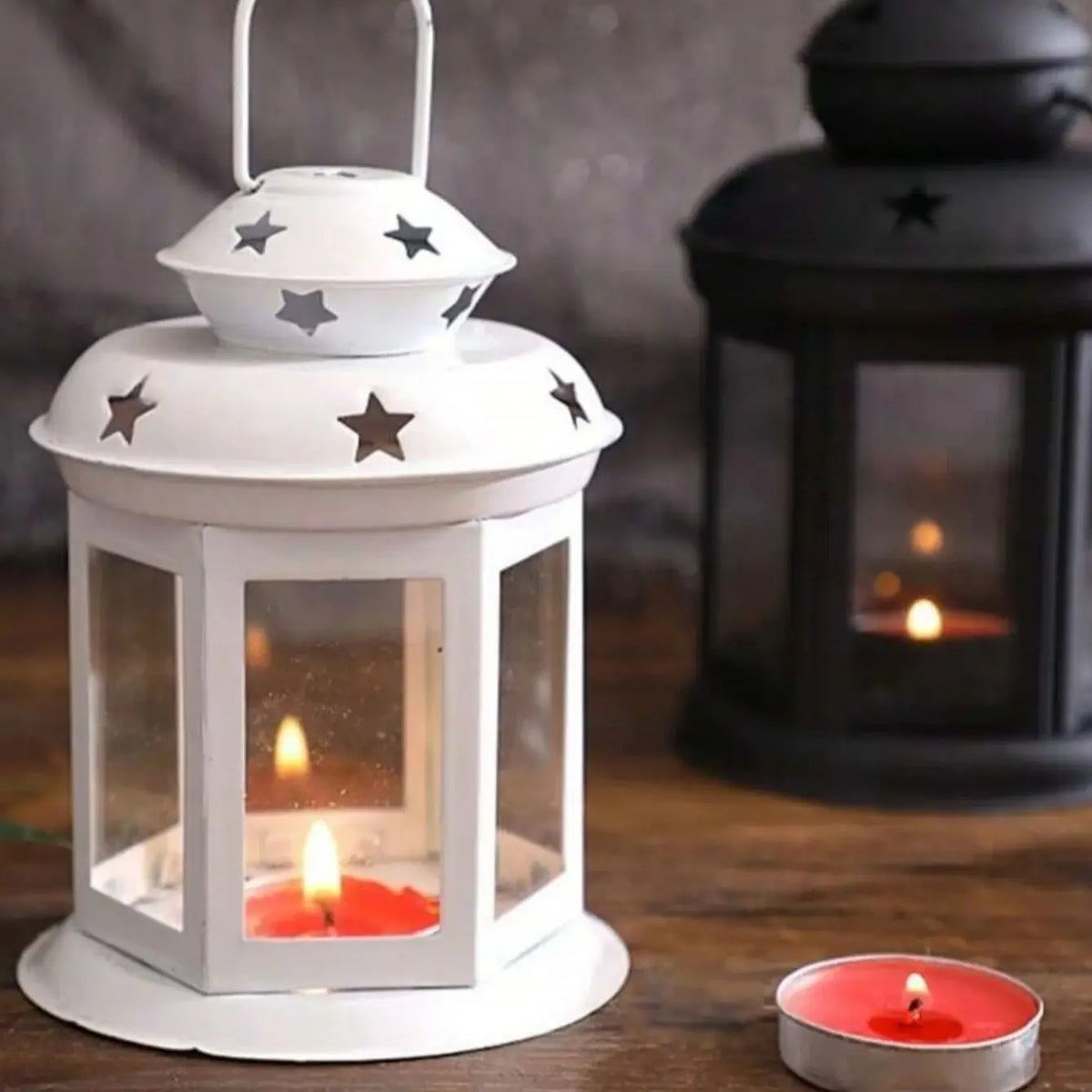 A white vintage hanging metal lantern with a glass enclosure and a lit candle on a wooden surface.