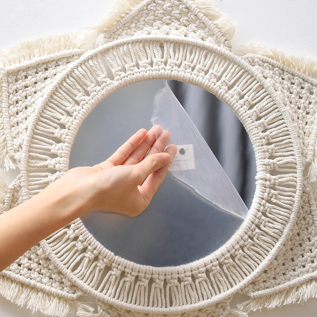 A hand is placing a piece of clear plastic onto a round macrame wall mirror for bohemian home decor.