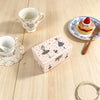 A pink ballet-themed music box with rotating figurines and jewelry storage sits on a wooden table next to a teacup and saucer