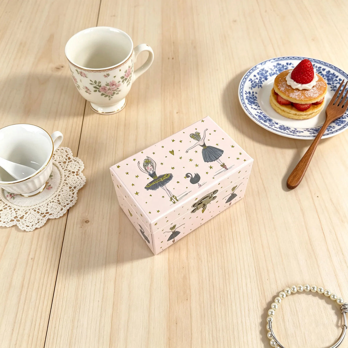 A pink box featuring ballet-themed illustrations sits on a wooden table next to a teacup and a plate with a pastry.