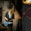 A bearded man in a dark shirt is working on metal tin signs in a dimly lit workshop.