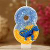 A birthday candle shaped like the number 8, featuring a blue glitter top and an orange base with a small blue dinosaur figure