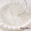 A clear glass magnifying glass is positioned over a white dish filled with small, round, clear spacer beads for jewelry makin