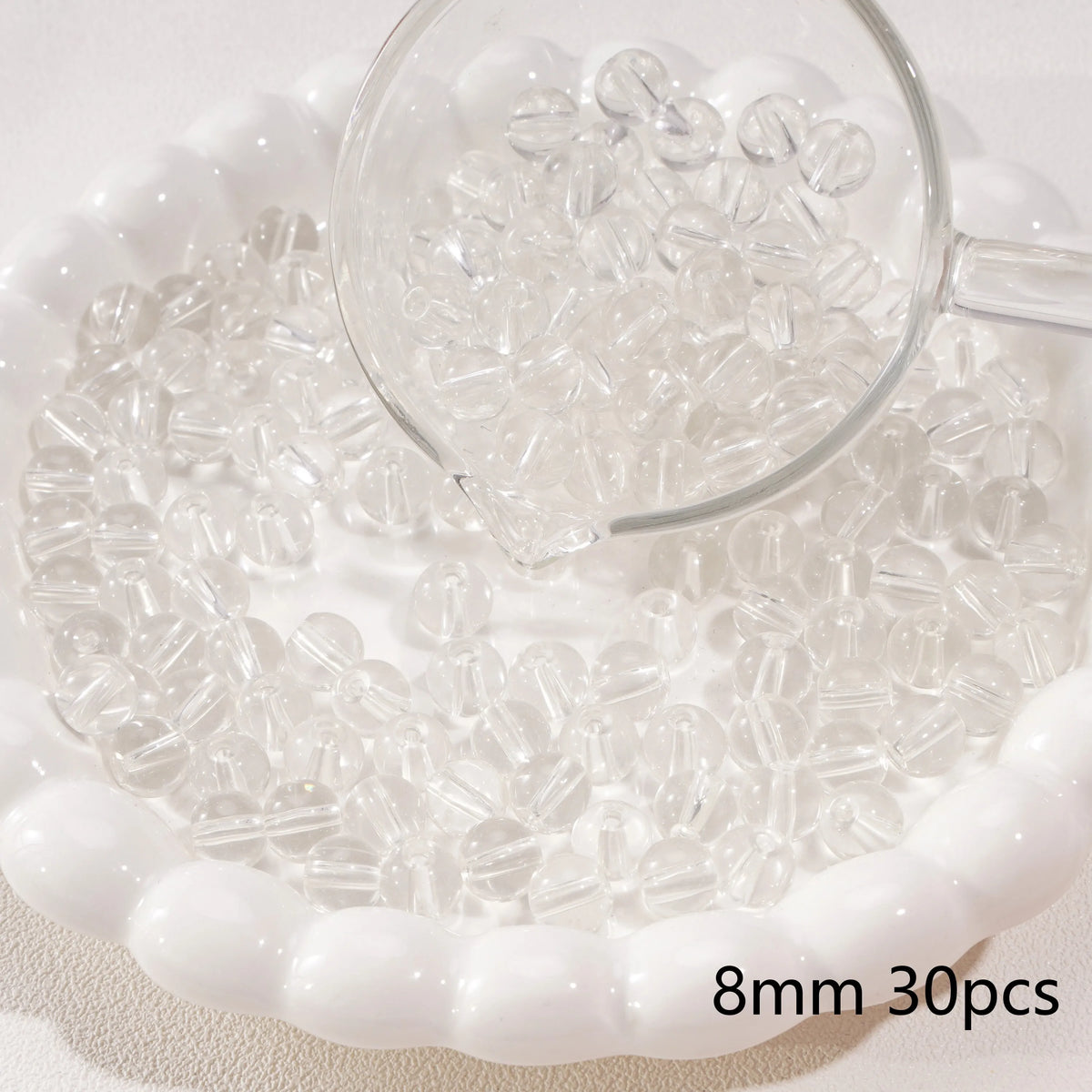 A clear glass magnifying glass is positioned over a white dish filled with small, round, clear spacer beads for jewelry makin