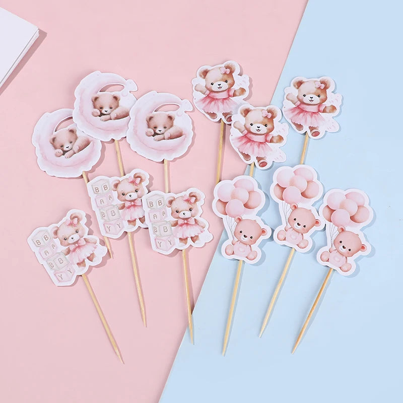 Pink cartoon bear cupcake toppers on wooden sticks arranged on pink and blue paper.