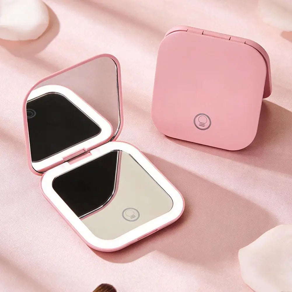 A pink portable folding LED makeup mirror with a black reflective surface is shown on a light pink background.