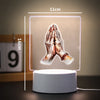 A 3D LED night light illuminates a pair of hands clasped in prayer against a soft background.