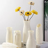 A white ceramic vase holds dried yellow and cream flowers against a light background.