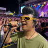 A young man wearing a yellow half-face latex mask performs on stage with a microphone under bright lights.