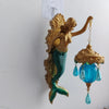 A vintage bronze sculpture of a mermaid with a green tail hangs on a white wall, featuring a blue glass lamp.