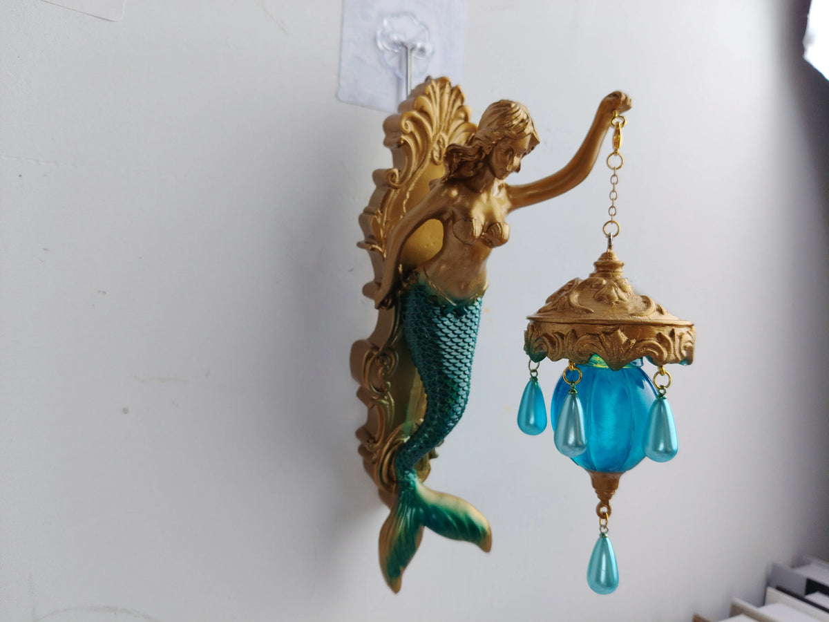 A vintage bronze sculpture of a mermaid with a green tail hangs on a white wall, featuring a blue glass lamp.