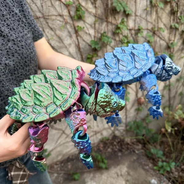 A hand holds a blue and green 3D printed alligator snapping turtle figurine with articulated joints against a blurred outdoor