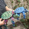 A hand holds a blue and green 3D printed alligator snapping turtle figurine with articulated joints against a blurred outdoor