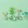 Three glass vials hold delicate pink and white floral arrangements on a light green background.