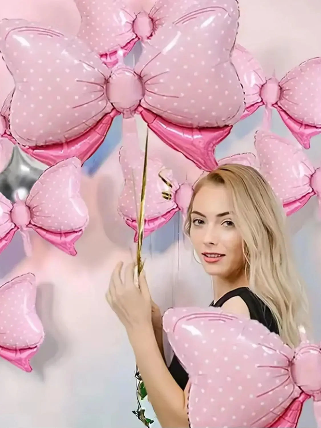 A woman is holding pink polka dot bow balloons for party decorations against a light background.
