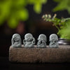 Four small gray stone monk statues arranged on a dark surface with blurred green foliage in the background.