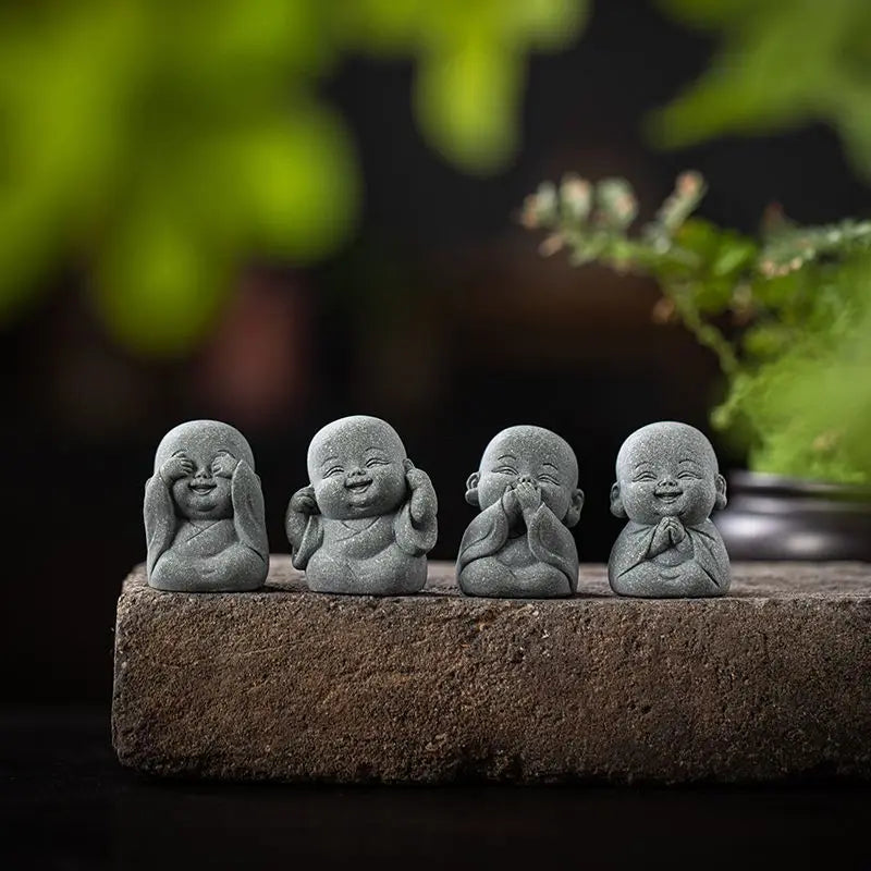 Four small gray stone monk statues arranged on a dark surface with blurred green foliage in the background.