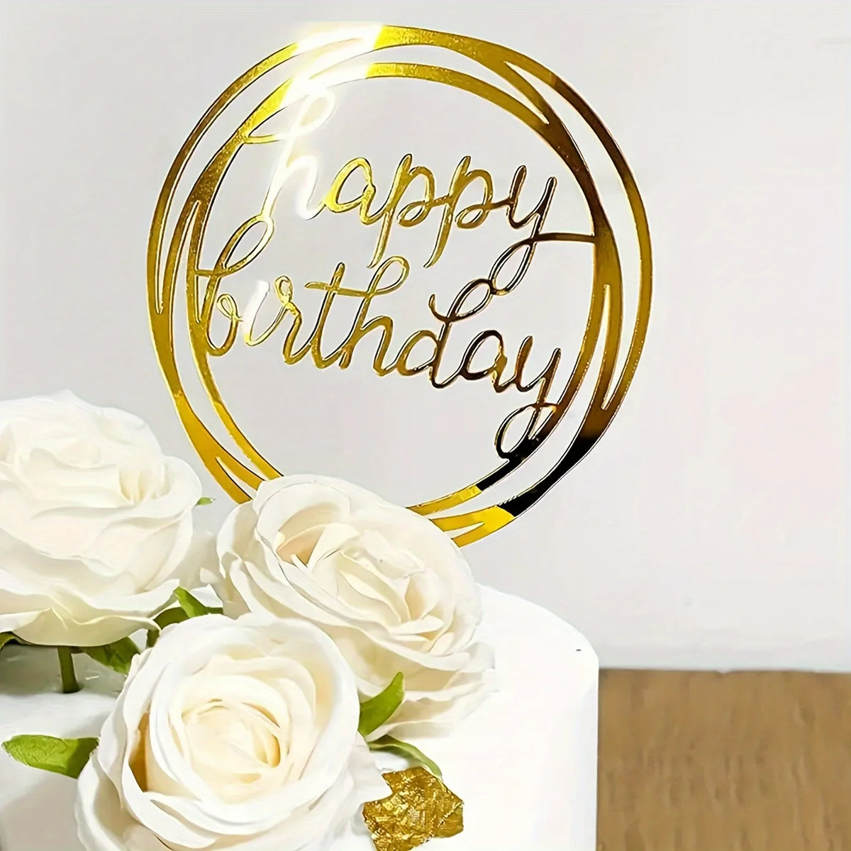 A gold acrylic 'Happy Birthday' cake topper is displayed next to white roses on a white cake.