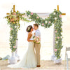 Un couple de mariés se tient sous une arche florale décorée de guirlandes d'eucalyptus pour une photo de mariage.