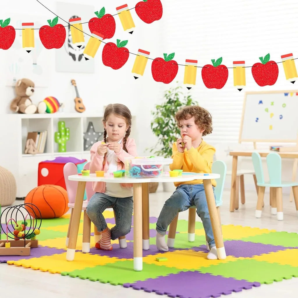 A colorful pencil and apple bunting banner strung across a room, with two young children sitting at a table.