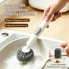 A hand is using a white long-handled liquid-adding rotating pot brush to wash dishes in a sink.