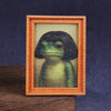 A framed illustration of a cute green frog in a wooden frame.