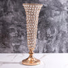 A tall, ornate crystal gold flower vase with a flared rim and foot stands on a white marble surface against a gray textured w