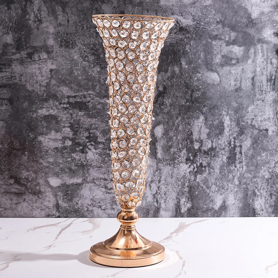 A tall, ornate crystal gold flower vase with a flared rim and foot stands on a white marble surface against a gray textured w