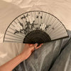 A folding hand fan with a gray and black striped design and white floral print is held by a person.