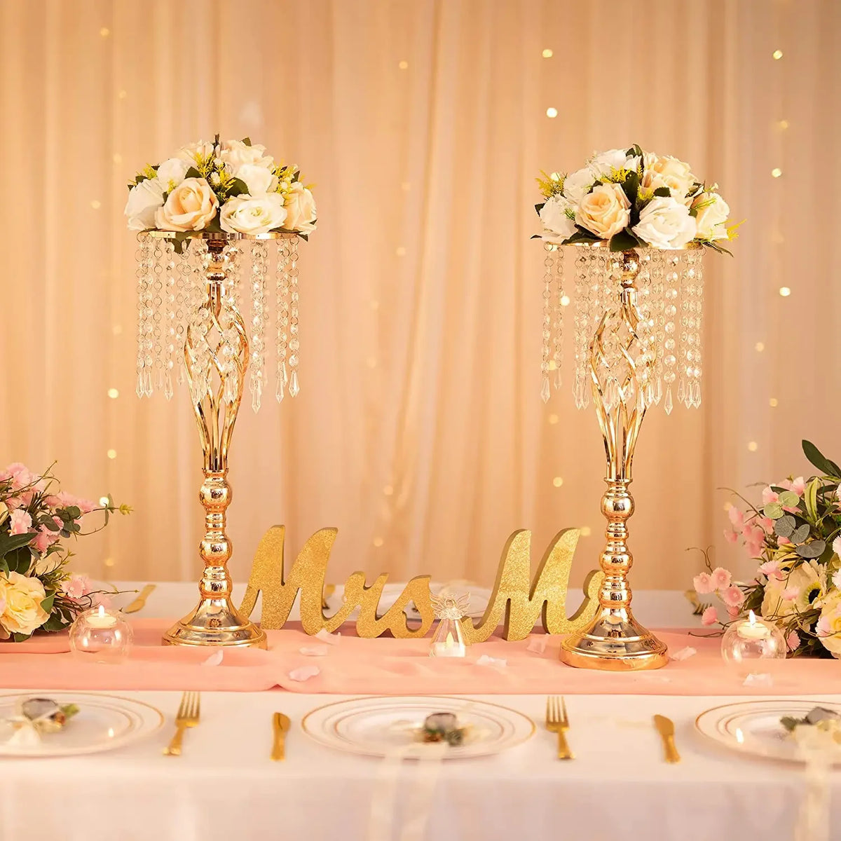 A set of gold crystal vases used as wedding centerpieces on a pink-draped table.
