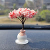 A small tree ornament with pink fluffy flowers sits on a white ceramic base on a dark dashboard surface.