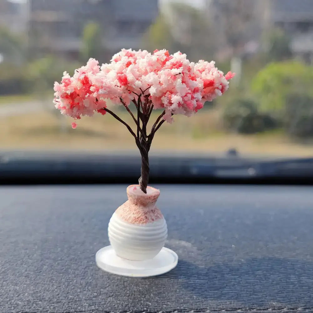 A small tree ornament with pink fluffy flowers sits on a white ceramic base on a dark dashboard surface.