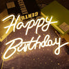 A glowing neon sign spelling out Happy Birthday is displayed on a dark surface with gold glitter and a black box in the backg