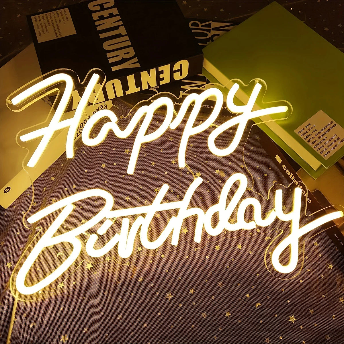 A glowing neon sign spelling out Happy Birthday is displayed on a dark surface with gold glitter and a black box in the backg