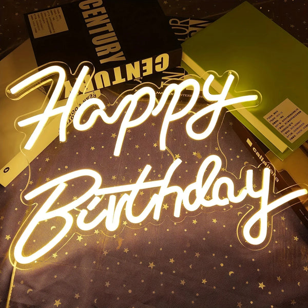 A glowing neon sign spelling out Happy Birthday is displayed on a dark surface with gold glitter and a black box in the backg
