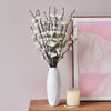 A vase filled with white artificial plum blossoms sits on a wooden surface next to a pink abstract framed print.